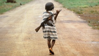 A child soldier holding a gun on their back walking down a path.